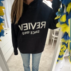 Svart hoodie med vit text från Review - Svart hoodie från Review med vit text 'REVIEW SINCE 1992' på bröstet. Tröjan har huva och en stor magficka framtill. Perfekt för en avslappnad stil.