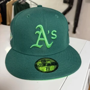 Grön Oakland Athletics keps från New Era - Snygg grön keps från New Era 59FIFTY med Oakland Athletics logga broderad framtill och patch på sidan. Klassisk flat peak och broderade detaljer. Perfekt för dig som gillar baseball eller vill ha en sportig stil.