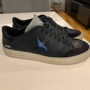 Axel Arigato Clean 90 Tripple bee Bird  - Säljer ett par svarta sneakers från 1,40MM med vit sula och blått broderi av en fågel på sidan. Skorna har snörning och detaljer i mocka vid tån. Perfekta för dig som vill ha en unik och stilren sneaker.