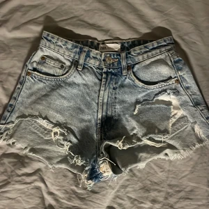 Ljusa slitna jeansshorts från Zara - Säljer ett par ljusblå jeansshorts från Zara med råa kanter och stora slitningar framtill. Klassisk femficksmodell med hög midja och knappgylf. Perfekta för sommaren och ger en cool, avslappnad look.