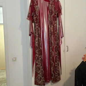 Vinröd långklänning med guldbroderi - Säljer en vinröd långklänning med glansigt tyg och vackra guldbroderade detaljer längs kanterna och ett bälte som kommer med den. Klänningen har lång ärm och en elegant, flytande passform. Perfekt för speciella tillfällen när du vill sticka ut.