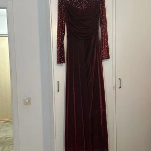 Vinröd glittrig långklänning med drapering - Säljer en vinröd långklänning med glittrigt tyg och draperad överdel. Klänningen har långa ärmar och en elegant släpdetalj. Perfekt för festliga tillfällen när du vill sticka ut.