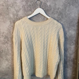 Beige stickad tröja med kabelmönster - Mysig beige stickad tröja med  kabelstickat mönster framtill och på ärmarna. Tröjan har rund halsringning och långa ärmar.