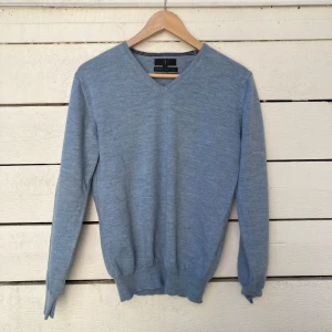 Ljusblå merinoullströja från Jasper Conran - Säljer en tunn och mjuk ljusblå tröja från Jasper Conran i exklusiv italiensk merinoull. Tröjan har v-ringning, långa ärmar och ribbade muddar. Perfekt för lager-på-lager och stilrena outfits.