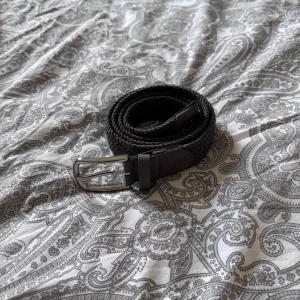 Svart flätat bälte med silverfärgat spänne - Säljer ett grått flätat bälte med silverfärgat metallspänne. Bältet har detaljer i skinn vid spännet och änden, och passar perfekt till jeans eller chinos. Stilren och enkel design som funkar till många outfits. 105cm