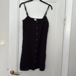 Svart knappklänning med smala axelband - Säljer en svart kortklänning med smala axelband och knappar framtill. Enkel och stilren design som passar perfekt till sommaren. Klänningen har en rak passform och är lätt att matcha med olika accessoarer.