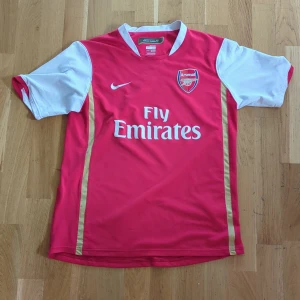 Arsenal hemmatröja Nike Fly Emirates - Säljer en klassisk Arsenal hemmatröja från Nike med Fly Emirates-tryck. Tröjan är röd med vita ärmar och detaljer i guld. Broderat Arsenal-märke på bröstet och Nike-logga. Tillverkad i lätt och ventilerande material.