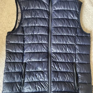 Blå dunväst från Original Matterhorn - Säljer en blå dunväst från The Jockey Maker med quiltad design och dragkedja framtill. Västen har två fickor med dragkedja och är perfekt för lager-på-lager-stil. Enkel och stilren modell som passar till många outfits.