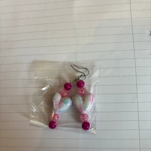 Handgjorda örhängen med hjärtan och pärlor - Säljer ett par unika örhängen med stora hjärtan i pastellfärger och glittriga rosa samt lila pärlor. Perfekta för att ge din outfit en färgglad och lekfull touch!