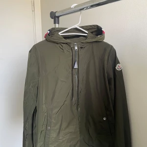 Moncler windbreaker 2st (vindjacka) med huva - Snygg vindjacka från Moncler med huva och dragkedja framtill. Jackan har Moncler-logga på ärmen och detaljer i rött, vitt och blått på huvan. Finns i både olivgrön och grå färg. Perfekt för blåsiga dagar.