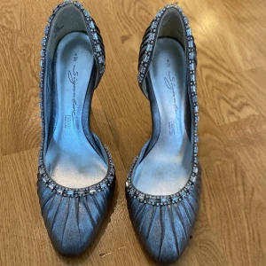 Silvriga pumps med kristaller - Eleganta silvriga pumps från next signature med glittrande kristaller längs kanten. Skorna har en rund tå och en smal klack som ger en sofistikerad look. Perfekta för speciella tillfällen.