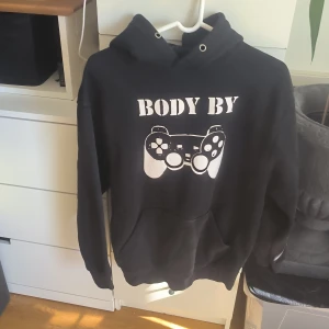 Svart hoodie från Tee Jays - Säljer en svart hoodie från Tee Jays med tryck av en spelkontroll och texten 'Body By'. Perfekt för gamingkvällar eller avslappnade dagar. Hoodien har en klassisk passform med känguruficka och justerbar huva.