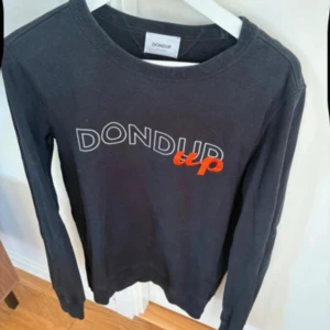 Svart långärmad tröja från Dondup - Säljer en svart långärmad tröja från Dondup med tryck i vitt och orange på bröstet. Tröjan har en rund halsringning och är perfekt för en avslappnad stil. Passar bra till jeans eller chinos.