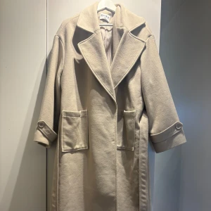 Beige kappa från NA-KD - Elegant beige kappa från NA-KD med stora slag och knappar på ärmsluten. Kappan har en klassisk design med stora fickor framtill och ett bälte baktill för en justerbar passform. Använd 1 gång. Som ny.  