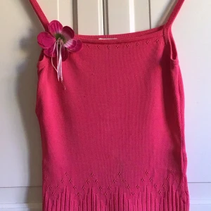 Rosa stickad linnetopp med blomdetalj - Säljer en söt rosa stickad linnetopp med smala axelband och en dekorativ blomma på axeln. Toppen har en fin fransdetalj längst ner som ger en lekfull touch. Perfekt för sommarens varma dagar!