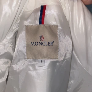 Vit dunjacka från Moncler - Säljer en stilren vit dunjacka från Moncler. Jackan har en quiltad design med huva och dragkedja. Perfekt för kyligare väder och ger en lyxig känsla. Inuti finns en unik serietidningsetikett som ger en rolig touch. Det går att scanna loggan vid armen.