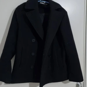 Svart kappa med knappar - Elegant svart kappa med stora knappar och bred krage. Perfekt för kyligare dagar med sin klassiska design och långa ärmar. En tidlös stil som passar till många tillfällen.