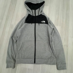 Grå/svarthoodie från The North Face - Säljer en snygg grå hoodie från The North Face med svart panel och dragkedja framtill. Den har en justerbar huva och logga på både fram- och baksidan. Perfekt för en avslappnad stil.
