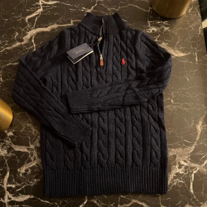 Mörkblå stickad tröja från Polo Ralph Lauren - Säljer en stilren mörkblå stickad tröja från Polo Ralph Lauren med kabelstickat mönster och en dragkedja vid halsen. Tröjan har en broderad röd logotyp på bröstet och långa ärmar. Perfekt för kyligare dagar!