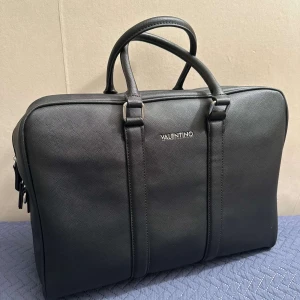 Svart portfölj från Valentino - Elegant svart portfölj från Valentino i syntetmaterial. Väskan har dubbla handtag och en avtagbar axelrem för flexibilitet. Den stängs med dragkedja och har flera innerfickor för organisering. Perfekt för att bära dina viktiga dokument och enheter med stil.