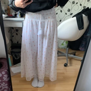 Vit långkjol med blommigt mönster - Säljer en vit långkjol med ett sött blommigt mönster i rosa och grönt från Gina Young. Kjolen har en elastisk midja med knytband och tofsar för extra stil. Perfekt för en sommaren! Det är en barn storlek 146/152 men passar som XS.