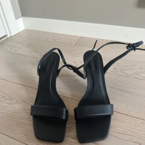 Svarta sandaler med klack - Eleganta svarta sandaler med klack och smala remmar. De har en fyrkantig tå och knytning runt ankeln för en stilren look.