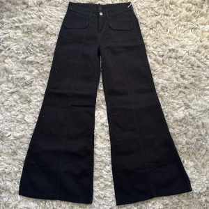 Svarta vida byxor med fickor - Snygga svarta byxor med låg midja och vida ben. Byxorna har framfickor med lock och en knappstängning i midjan. Perfekta för en stilren look. Aldrig använda, prislappen kvar 