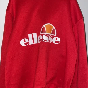 Röd hoodie från Ellesse - Säljer en snygg röd hoodie från Ellesse med deras ikoniska logga i vitt och orange på framsidan. Tröjan har en klassisk huva och långa ärmar, perfekt för en avslappnad stil.