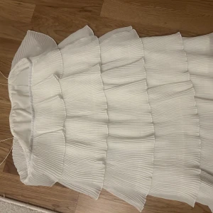 Vit axelbandslös klänning - Säljer en elegant vit  klänning med volanger. Perfekt för sommarens fester. Klänningen har en luftig och lätt design som ger en romantisk känsla. Passar utmärkt för en stilren look.