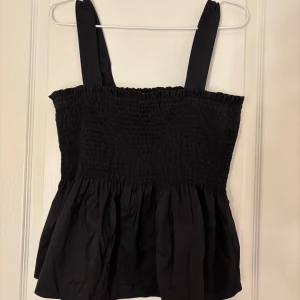 Svart linnetopp från Vintage Stories - Säljer en svart linnetopp från Vintage Stories med smockdetaljer och breda axelband. Toppen har en luftig och bekväm passform, perfekt för varma dagar. Passar bra till både jeans och kjol. Banden går att vika ner så den blir till en tubtopp.