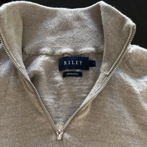 Beige merinoulltröja från Riley - Säljer en stilren beige tröja i merinoull från Riley. Tröjan har en dragkedja vid halsen och långa ärmar, perfekt för kyligare dagar. Den är mjuk och bekväm, idealisk för både vardag och mer uppklädda tillfällen.