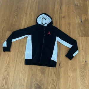 Svart och vit Jordan hoodie med dragkedja - Säljer en snygg svart hoodie med vita detaljer och dragkedja från Air Jordan. Den har en röd logga på bröstet och text på huvan. Perfekt för alla årstider och vid funderingar så är det bara att höra av sig! 