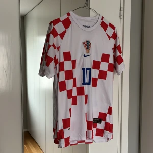 Kroatien fotbollströja från Nike - Säljer en vit och röd fotbollströja från Nike med Kroatiens landslagsemblem och nummer 10. Tröjan har ett klassiskt rutmönster och korta ärmar. Perfekt för fotbollsfans!