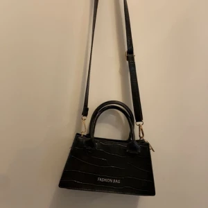 Svart axelväska med krokodilmönster - Snygg svart axelväska med krokodilmönster och texten 'FASHION BAG' på framsidan. Väskan har både handtag och en justerbar axelrem för flexibel användning. Perfekt för att ge en stilren touch till din outfit. Den är i ny skick knappt använd från Shein