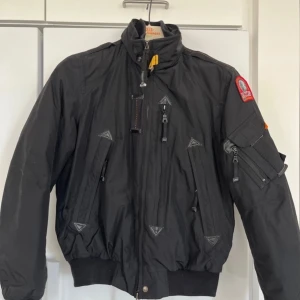 Svart bomberjacka från Parajumpers - Säljer en svart bomberjacka från Parajumpers med orange foder och flera praktiska fickor. Jackan har en dragkedja framtill och en broderad patch på ärmen. 