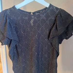 Svart blus från Holly & Whyte - Säljer en elegant svart blus från Holly & Whyte med ett diskret rutmönster. Blusen har korta ärmar med volangdetaljer som ger en fin touch. Perfekt för att lyfta vilken outfit som helst!