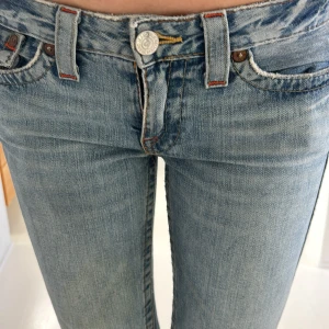 Blå lowaist bootcut jeans från True Religion - Snygga blå lowaist bootcut jeans från True Religion med orange/röd. Lite slitet vid bena men kolla bilderna för de andra defekter/slitningar. Innerbenslängd ca 83cm. Skriv om du undrar något!