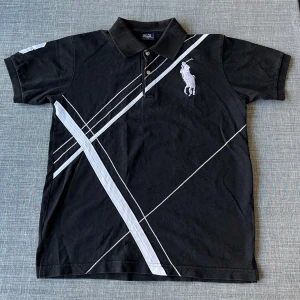 Svart pikétröja från Ralph Lauren - 📦Säljer en snygg svart pikétröja från Ralph Lauren med vita diagonala ränder och den ikoniska loggan på bröstet. Tröjan har korta ärmar och en klassisk krage med knappar. Perfekt för en stilren look! Storlek M, normal i storleken. Vid frågor, tveka inte att höra av er! ☺️