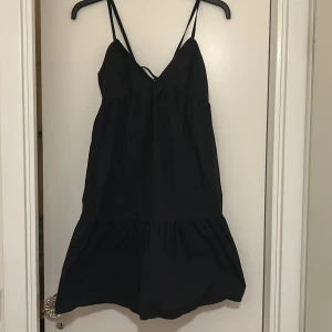 Svart volangklänning från BikBok - Snygg svart klänning från BikBok med smala axelband och knytning i ryggen. Perfekt för en stilren look. Klänningen har en volangdetalj nedtill som ger en fin touch.