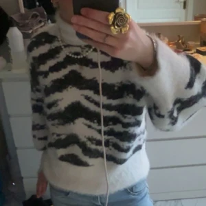 Vit och svart mönstrad stickad tröja - Säljer en mysig vit och svart stickad tröja med zebramönster. Tröjan har långa ärmar och en hög krage som håller dig varm under kyliga dagar. Perfekt för en stilren och bekväm look.