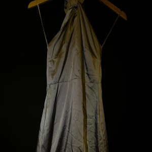 Grå långklänning med slits - Elegant grå långklänning i ett glansigt material med smala axelband och djup urringning. Klänningen har en stilren slits framtill som ger en sofistikerad touch.