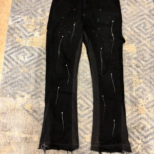 Svarta byxor från Gallery Dept. - Unika svarta byxor från Gallery Dept. med coola färgstänk och vita linjer som ger en konstnärlig touch. Byxorna har en loose passform och flera fickor för en avslappnad stil. Perfekta för dig som vill sticka ut med en kreativ look.