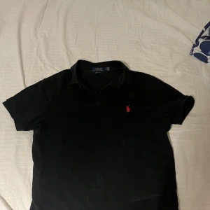 Svart pikétröja från Ralph Lauren - Säljer en klassisk svart pikétröja från Ralph Lauren med röd broderad logga på bröstet. Tröjan har korta ärmar och en stilren krage. Perfekt för en avslappnad men ändå stilfull look. Riktigt bra passform.