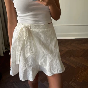Vit omlottkjol från H&M - Säljer en söt vit omlottkjol från H&M med knytband i midjan. Kjolen har ett fint broderat mönster och volangdetaljer längs nederkanten. Perfekt för varma dagar!