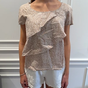 Beige blus med volangdetalj - Säljer en söt beige blus med ett diskret prickigt mönster och volangdetalj framtill. Blusen har korta ärmar och en rund halsringning. Perfekt för en stilren och chic look.