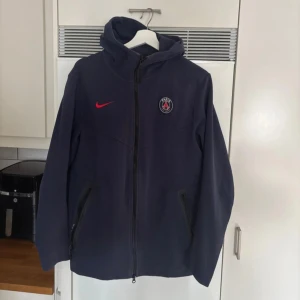  Nike PSG hoodie - Säljer en mörkblå hoodie från Nike med PSG loggan på bröstet som är köpt i Frankrike, Paris. Hoodien har en dragkedja framtill och två sidofickor med dragkedjor. Perfekt för sportiga tillfällen eller som en stilren vardagshoodie.