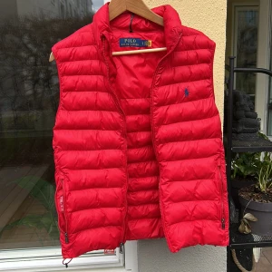 Röd dunväst från Polo Ralph Lauren - Säljer en snygg röd dunväst från Polo Ralph Lauren. Västen har en dragkedja framtill och är quiltad för extra värme. Perfekt nu till våren. Har en fläck framifrån därifrån lägre pris 