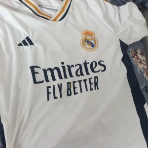 Vit Real Madrid fotbollströja - Säljer en vit Real Madrid fotbollströja från Adidas med korta ärmar. Tröjan har klubbens emblem och sponsorlogga på framsidan samt spelarnamnet Bellingham och nummer 5 på baksidan. Den har även guld- och svarta detaljer på kragen och ärmarna.