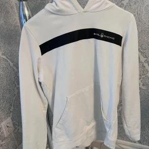 Vit hoodie från Sail Racing - Säljer en stilren vit hoodie från Sail Racing med en svart detalj över bröstet där loggan är tryckt. Tröjan har en klassisk känguruficka och en bekväm huva. Perfekt för en avslappnad stil. Sprickor i trycket, se bild