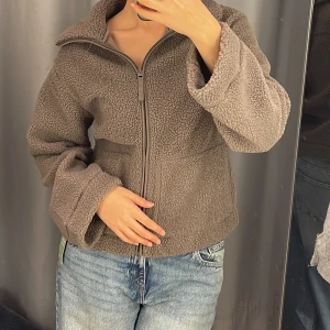 Brun teddyjacka med dragkedja - Mysig brun teddyjacka med dragkedja framtill. Jackan har en avslappnad passform och långa ärmar, perfekt för kyliga dagar. Den är gjord i ett mjukt och fluffigt material som håller dig varm och bekväm.